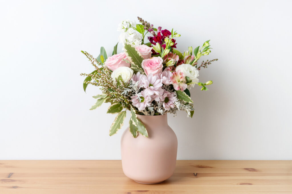 A vase with beautiful flowers on a wooden shelf again a white wall.